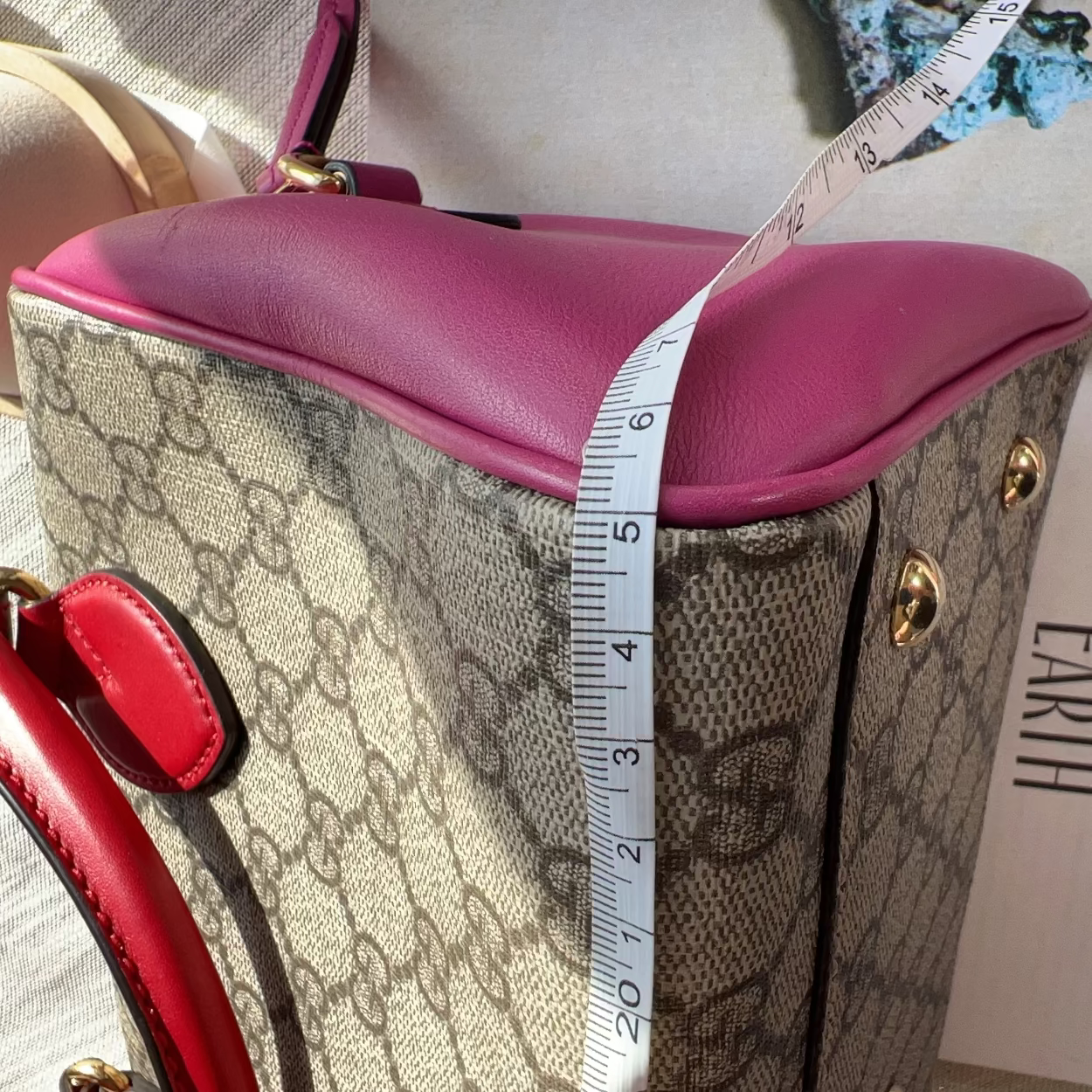Designer handbag with pink top and red handles, featuring a measuring tape for scale, on a surface with text in the background.