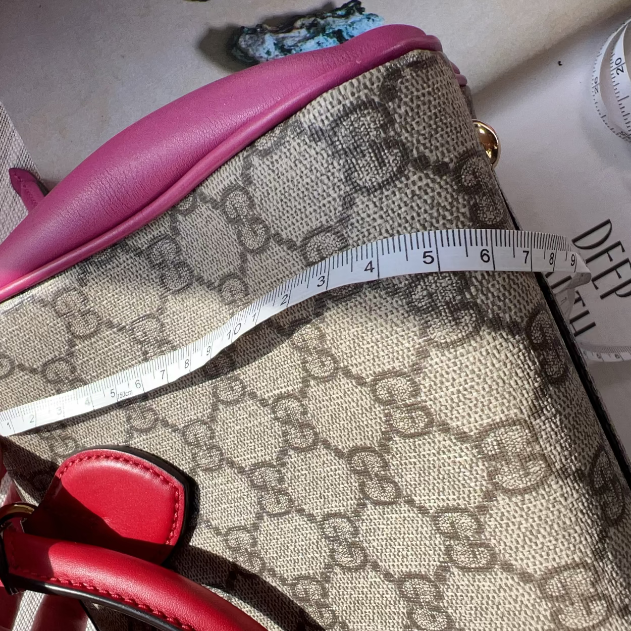 Handbag with pink top and red bottom section, featuring a pattern, held by a hand with a measuring tape on top.