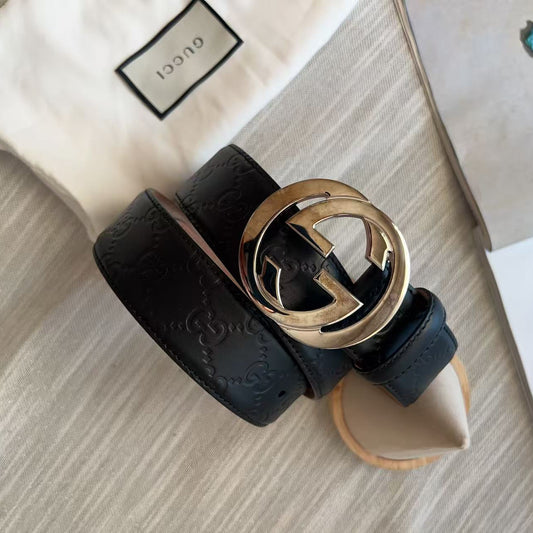 A black Gucci leather belt with the brand's signature interlocking G buckle, coiled on a light beige textured surface next to its cream Gucci dust bag.