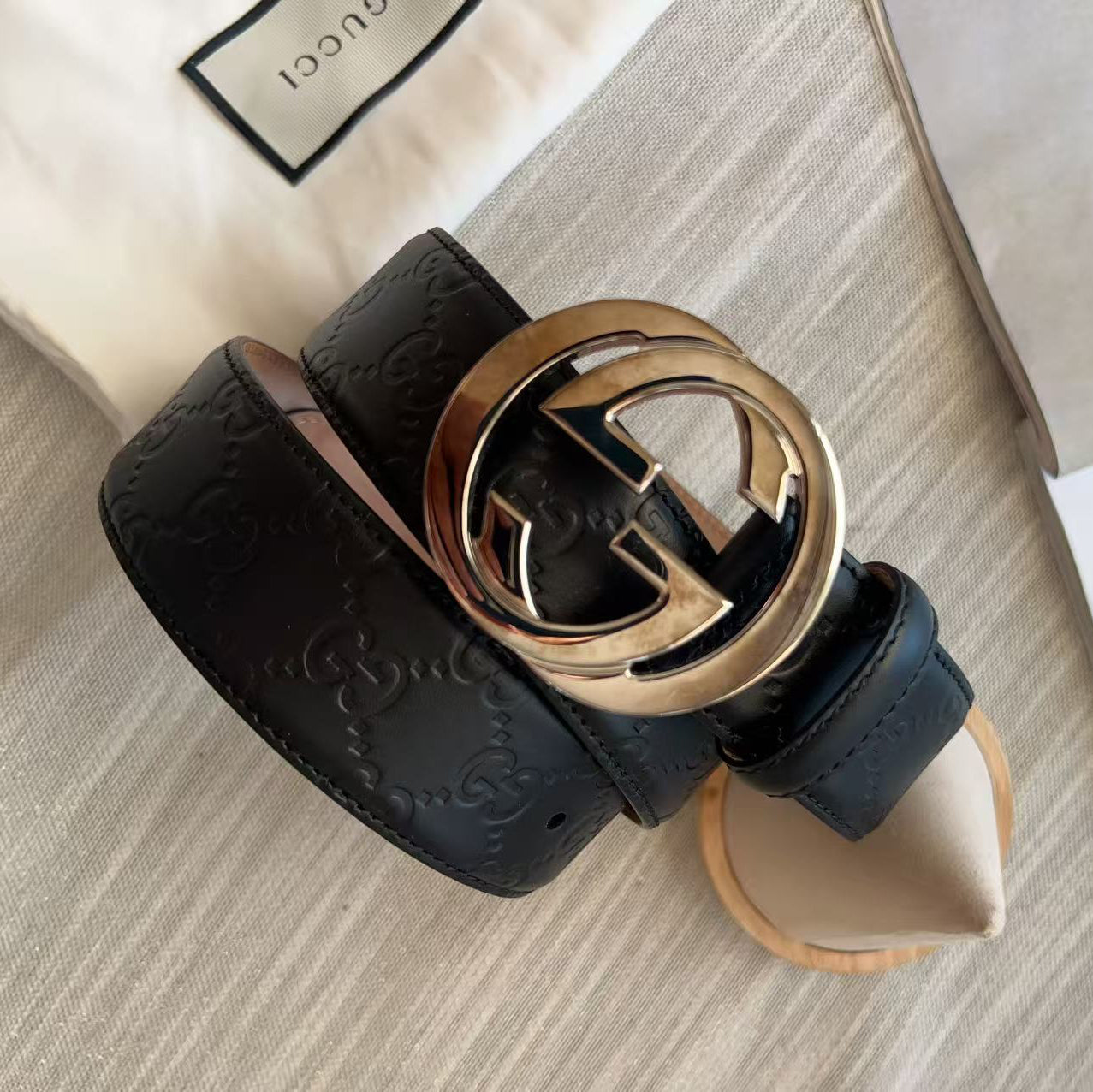 A black leather belt with an embossed monogram pattern and a large gold-tone interlocking G buckle, shown coiled on a light beige surface with a Gucci dust bag and printed papers nearby.