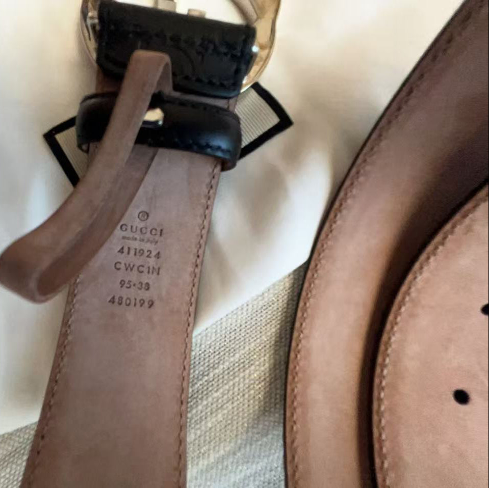 A close-up of a Gucci belt showing the interior stamp and a brown leather strap with a black leather keeper and gold-tone metal ring, laid on a white fabric background.