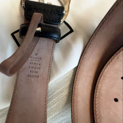 A close-up of a Gucci belt showing the interior stamp and a brown leather strap with a black leather keeper and gold-tone metal ring, laid on a white fabric background.