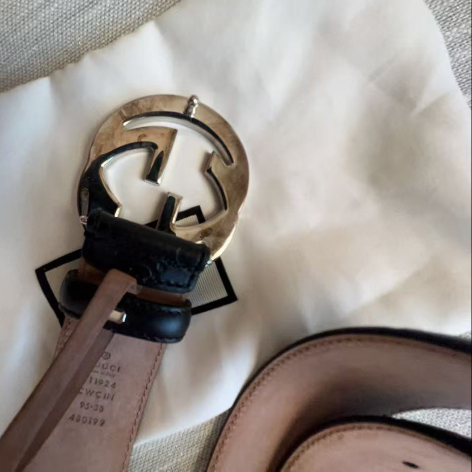 A close-up of a Gucci belt with a signature interlocking G buckle, resting on a white Gucci dust bag. The belt features a light brown leather strap with black leather accents and a gold-tone buckle.