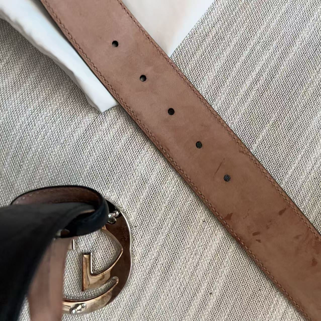 A close-up of a brown leather strap, likely a belt or bag strap, laid diagonally on a textured beige and white striped fabric surface. A gold-tone metal buckle is partially visible in the bottom-left corner.