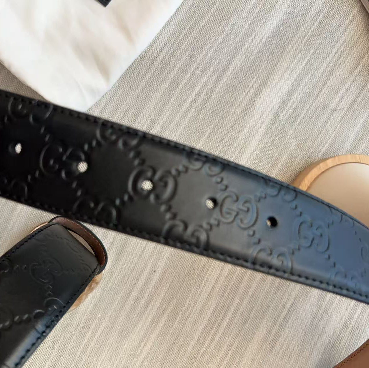 A close-up of a black leather belt with an embossed interlocking GG monogram pattern, held by a hand over a light beige textured fabric background.