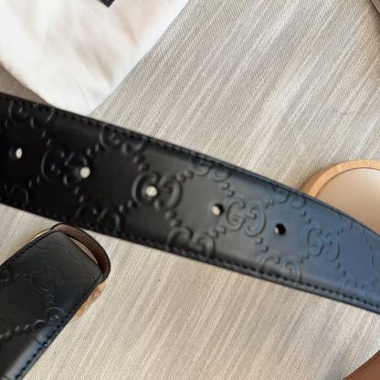 A close-up of a black leather belt with an embossed interlocking GG monogram pattern, held by a hand over a light beige textured fabric background.