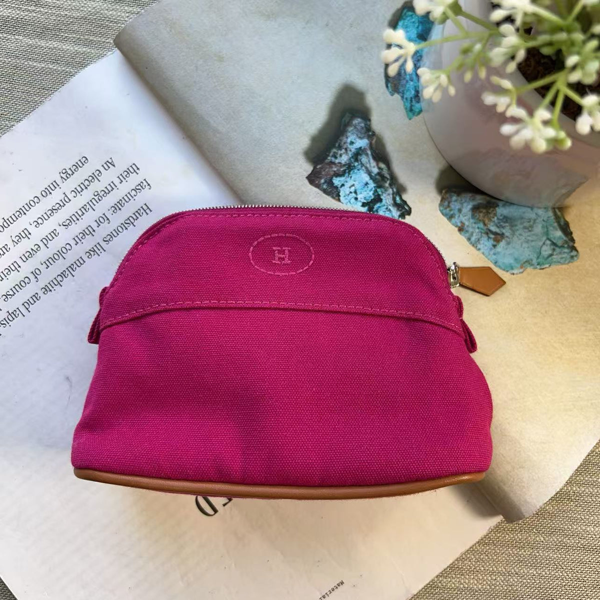 A bright pink canvas zippered pouch with a brown leather trim and a circular embroidered 'H' logo, styled on an open book with decorative stones and small white flowers.
