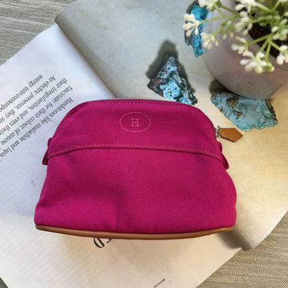 A bright pink canvas zippered pouch with a brown leather trim and a circular embroidered 'H' logo, styled on an open book with decorative stones and small white flowers.