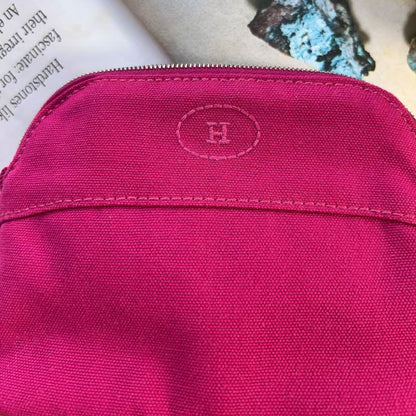 A close-up of a bright pink canvas pouch with a zippered top and an embroidered oval logo featuring the letter 'H' on the front pocket. The pouch is styled on a light beige surface next to a white book page and textured stones.