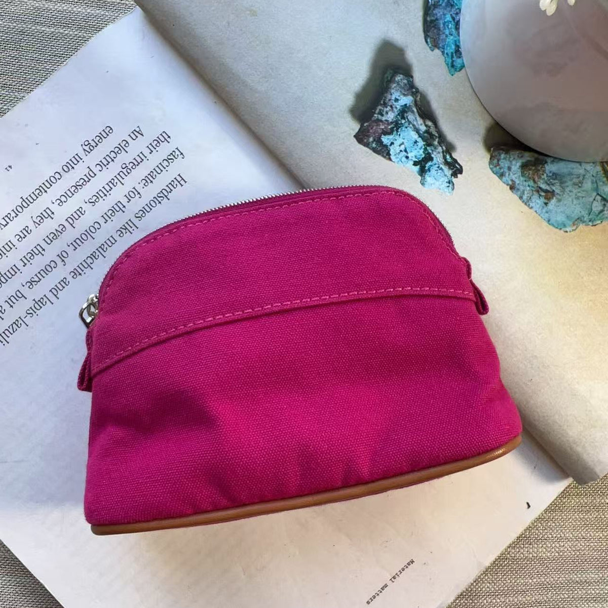 A bright magenta pink fabric pouch with a curved top and brown leather trim, resting on an open book with decorative stones and a ceramic pot in the background.