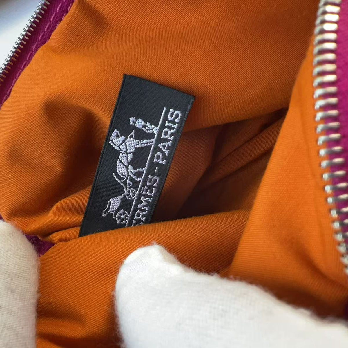 Close-up of an orange Hermès Paris pouch or bag interior with a black woven brand label, silver zipper, and white knit fabric at the bottom.