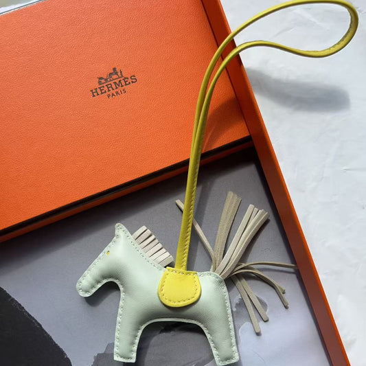 An Hermes Pegasus charm in pale sage green and yellow leather, displayed inside its orange Hermes box with its signature strap looped above.