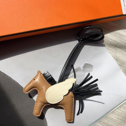 A small brown leather winged horse charm with black tinsel-like wings and a cream wing, laid on a white surface next to an orange box. No person or real environment is present, so this is Product/Packaging rather than Lifestyle.