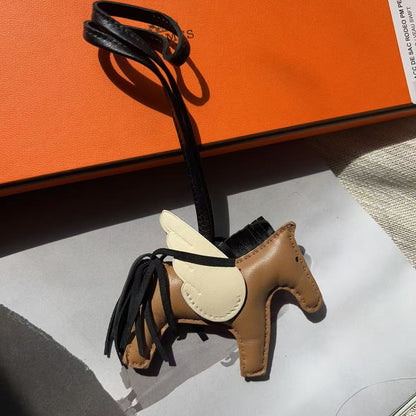 A miniature brown leather horse-shaped charm with black tassels and a black loop, shown next to an orange box on a white surface.
