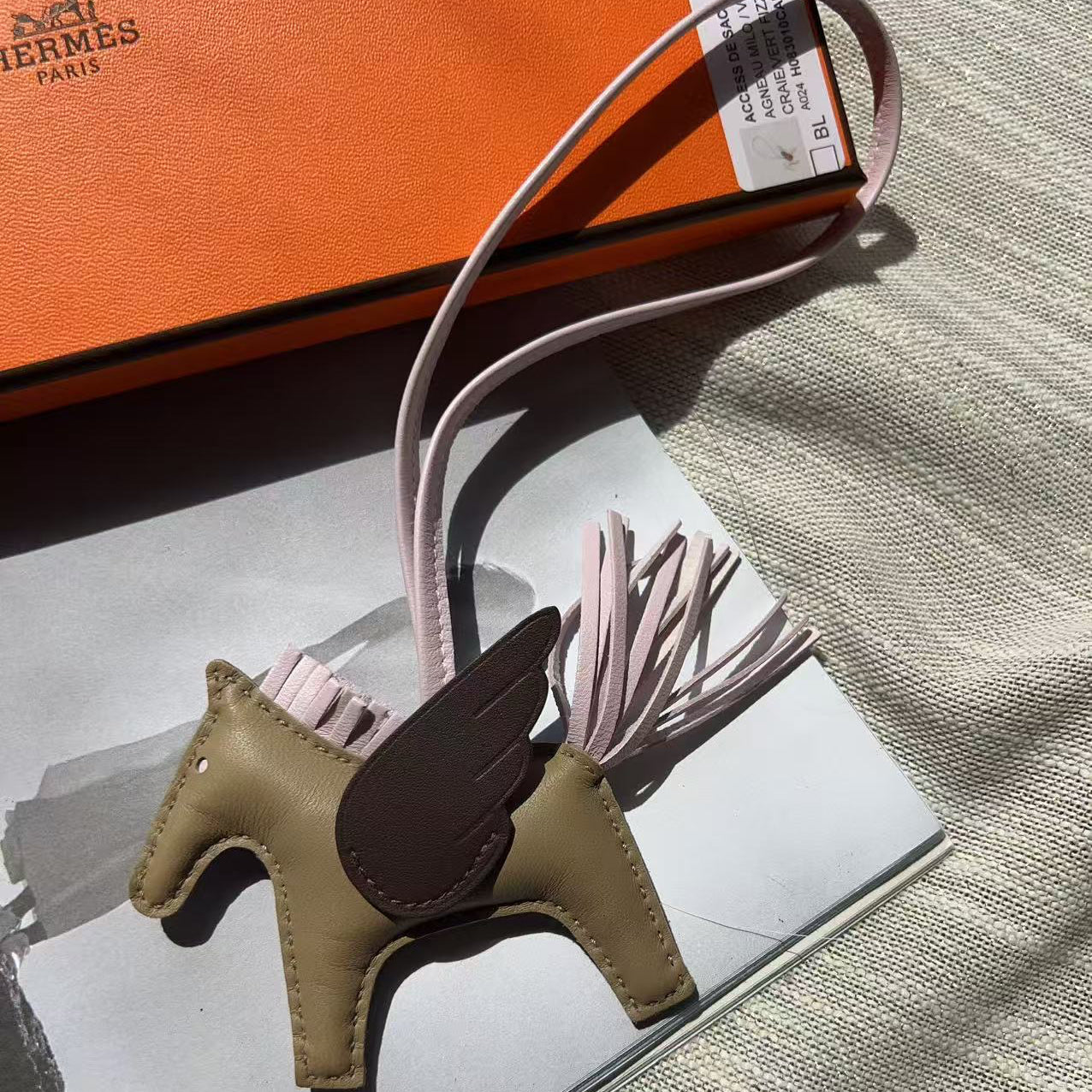 An Hermes winged horse charm in tan and dark brown leather with pale pink tassel straps, displayed on a silver tray next to an orange Hermes box.
