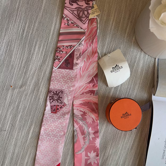 Pink patterned scarf with a red border on a textured surface next to a white box and orange container.