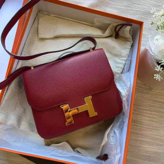 A burgundy pebbled leather handbag with a gold H-shaped turn-lock closure, presented inside an orange and white box with a beige dust bag.