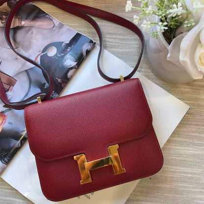 A red textured leather handbag with a large gold H-shaped clasp, styled on top of a white book with a magazine and small flowers in the background.