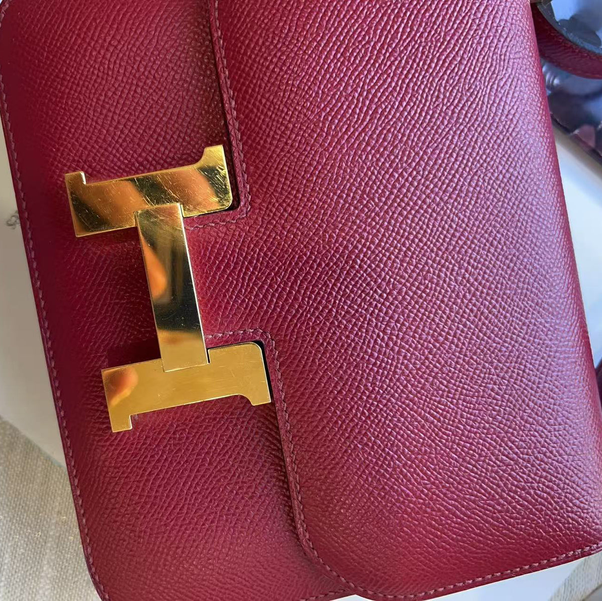 A close-up of a red textured leather handbag featuring a prominent polished gold-tone H-shaped metal clasp, resting on a light beige textured surface.