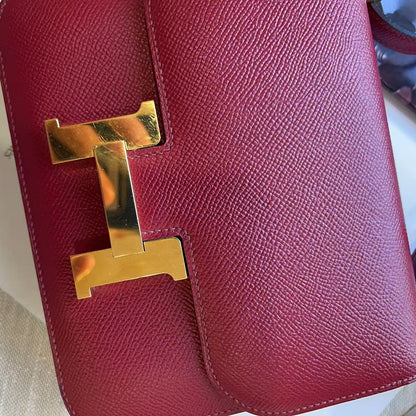 A close-up of a red textured leather handbag featuring a prominent polished gold-tone H-shaped metal clasp, resting on a light beige textured surface.