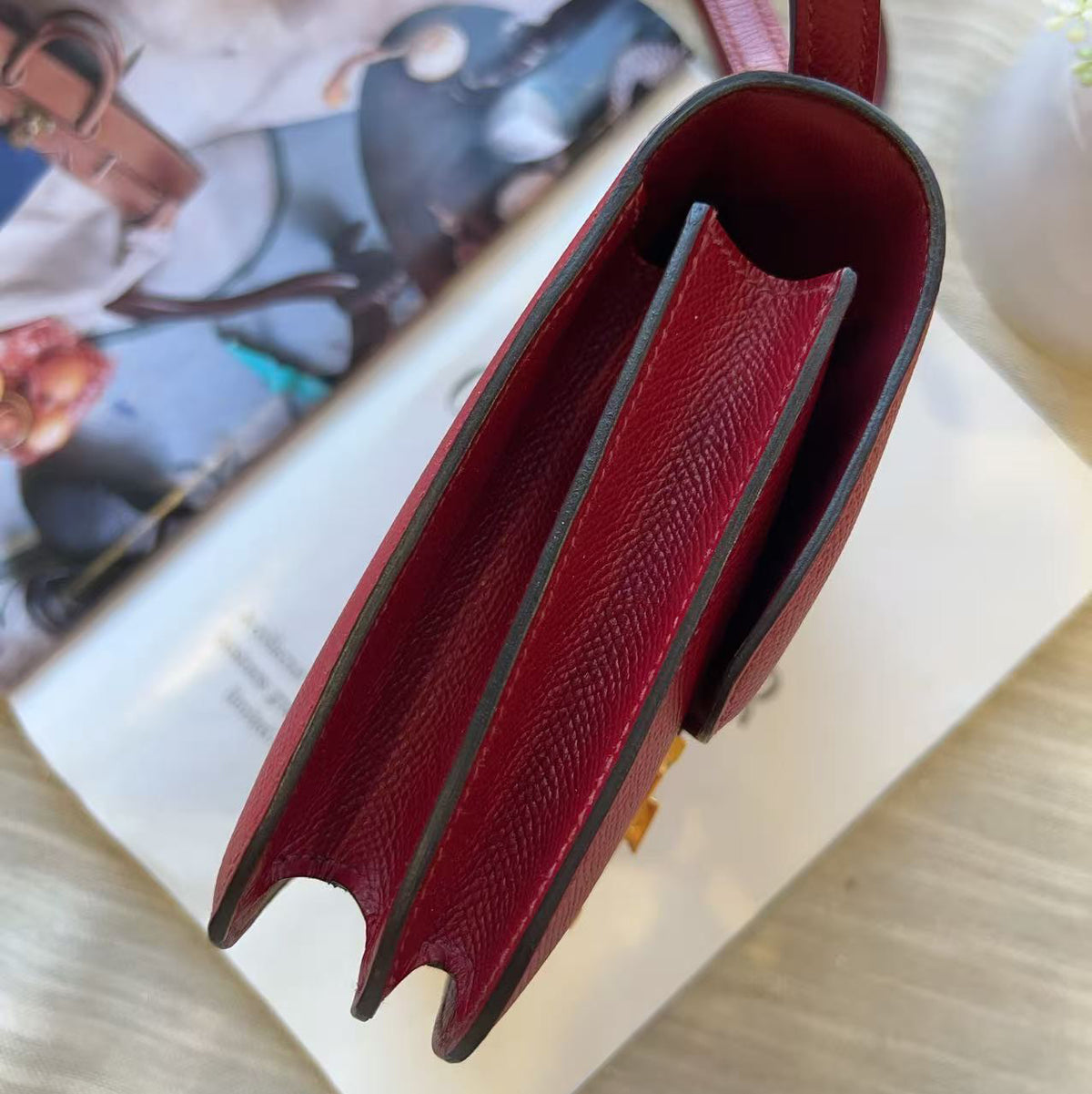 A close-up of a burgundy textured leather handbag, showing the interior lining and strap, styled on a light wood surface with a magazine in the background.