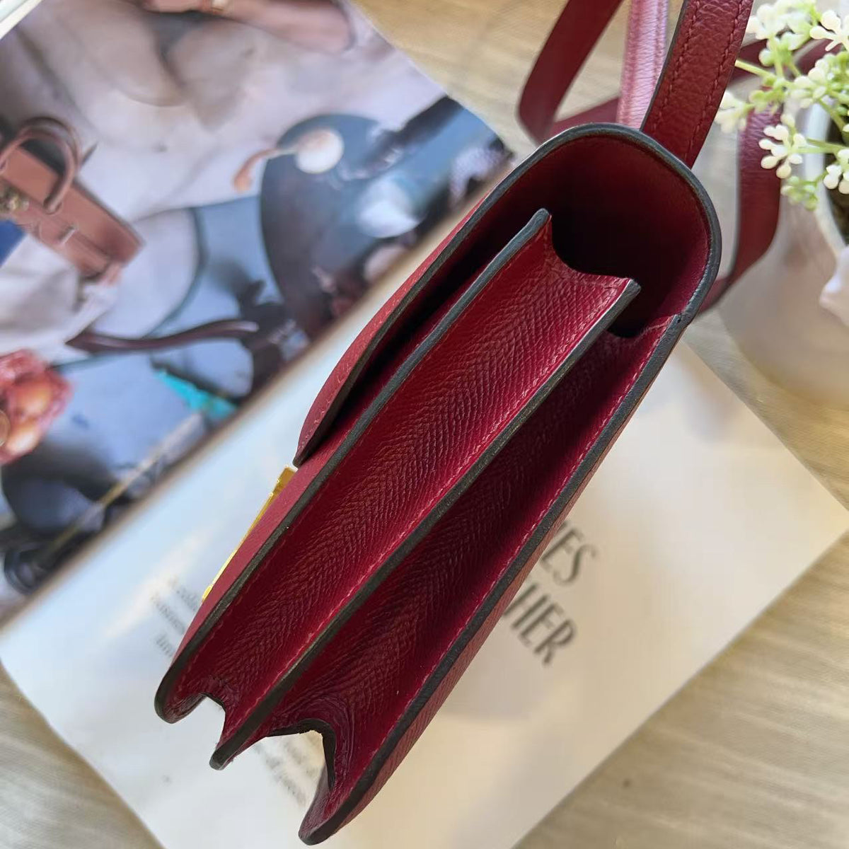 A close-up of a burgundy textured leather handbag, showing the interior lining and strap, resting on a light-colored wooden surface next to a magazine.