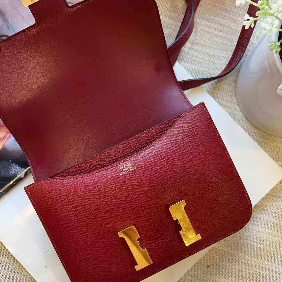 A red textured leather handbag shown open, with gold-tone hardware and a matching red strap, photographed on a light fabric surface with a white card underneath.