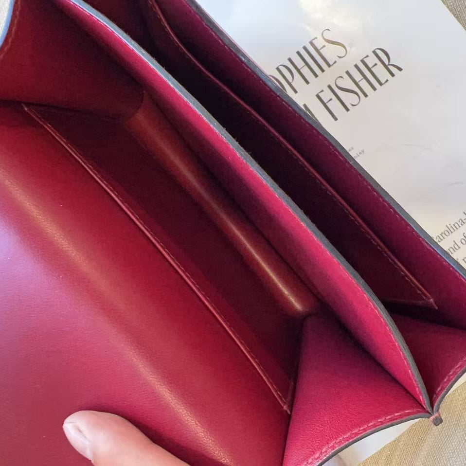 Close-up of an open burgundy leather clutch or wallet showing the interior lining and card slots, with fingers holding it open and a book visible in the background.