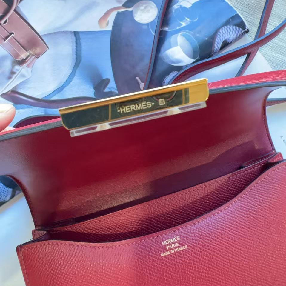A close-up of a Hermès red leather handbag showing the interior flap, gold-toned hardware with the Hermès logo, and part of the strap, with a magazine page underneath.