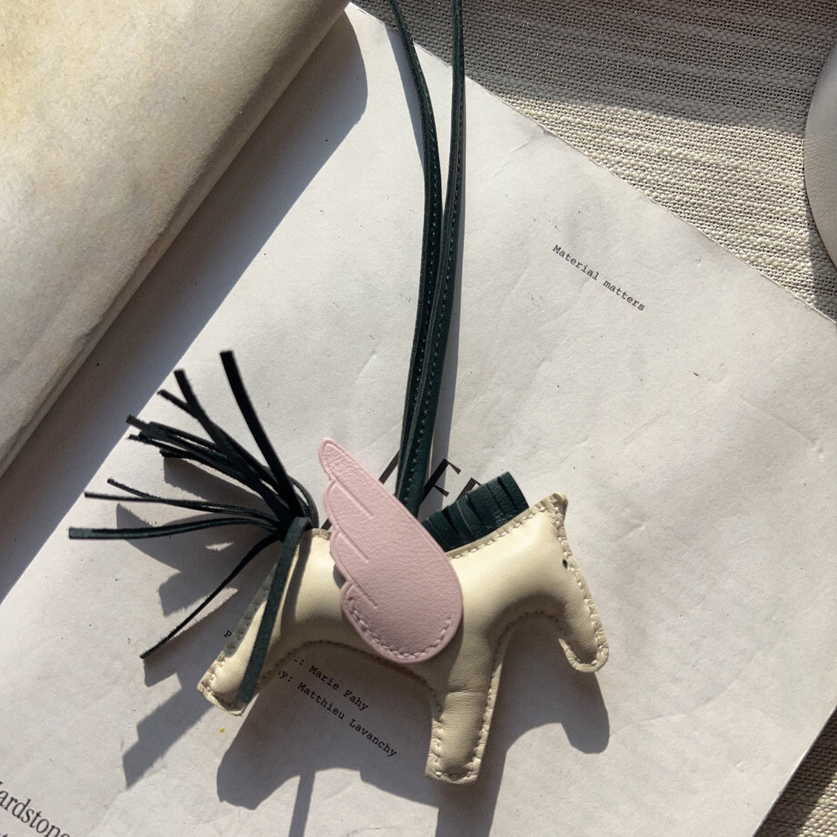 A small leather horse-shaped charm or bookmark with a pale pink wing, dark green strap, and black tassel, resting on an open book.
