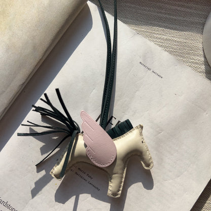 A small leather horse-shaped charm or bookmark with a pale pink wing, dark green strap, and black tassel, resting on an open book.