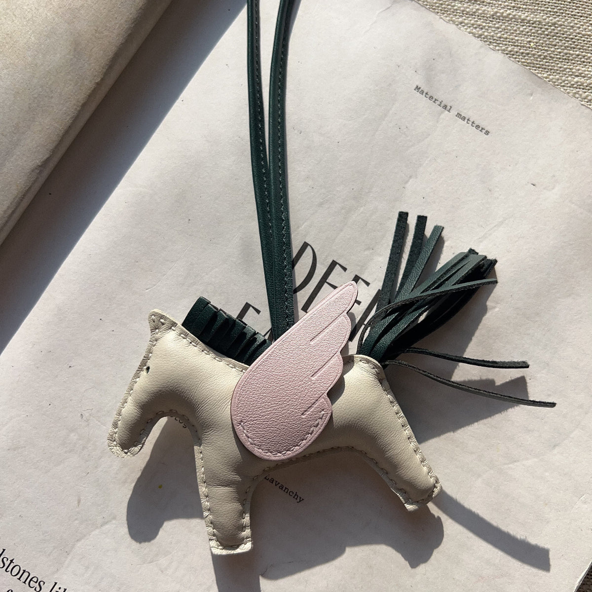 A small handmade leather horse-shaped charm or bag charm in off-white with a pale pink wing detail and dark green tassel/strap, photographed on top of an open book.