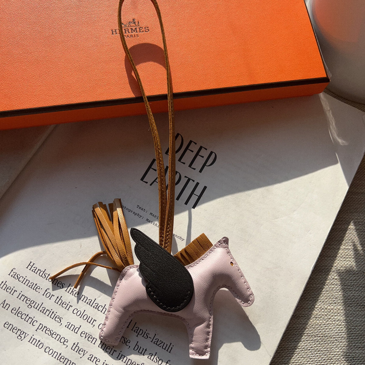 A small pink horse-shaped leather charm with a black wing and tan strap, resting on an open book with an orange Hermes box in the background.