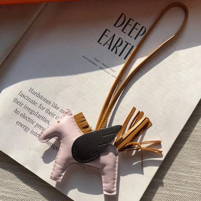 A light pink leather winged horse charm with a black wing and tan strap, resting on a white book page with a textured beige surface underneath.