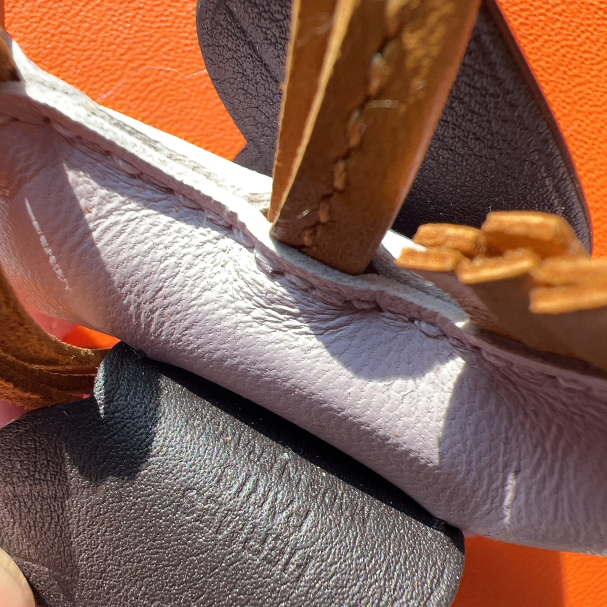 Close-up detail of a leather handbag showing a light beige textured leather strap, a dark gray leather accent piece, and a tan leather handle against a bright orange background.