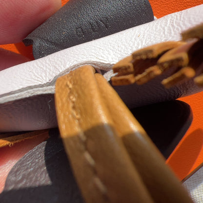 Close-up detail of a hand holding a white leather item with a dark gray leather tab and tan leather strap against an orange textured background.