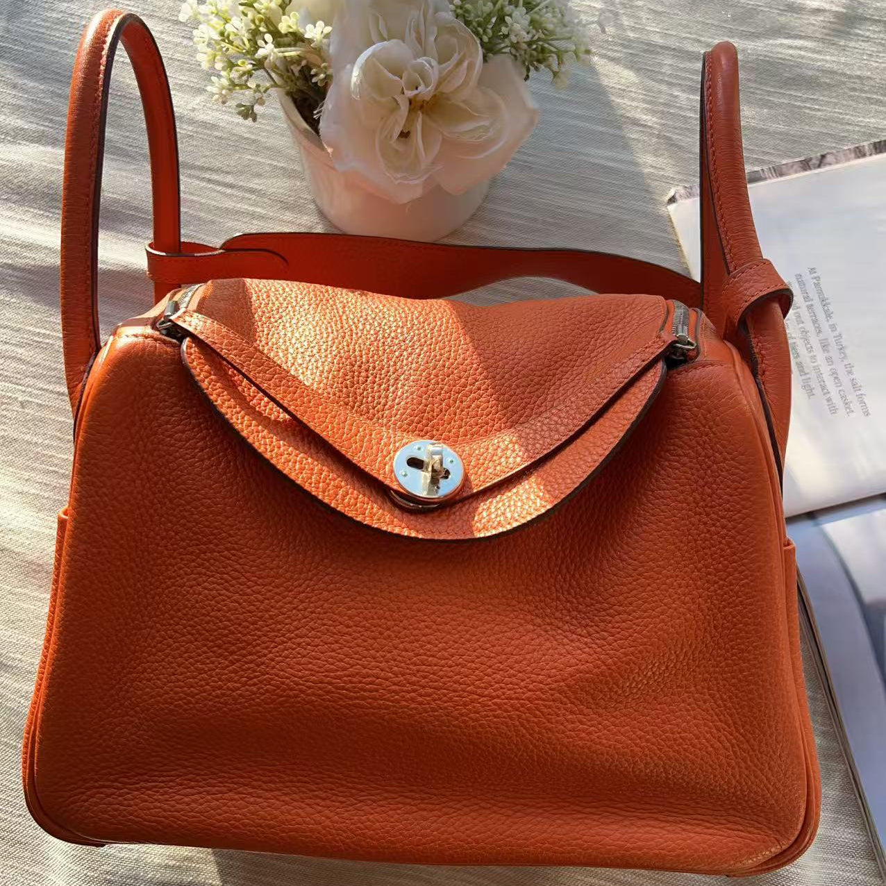 Orange handbag on a wooden surface with flowers and a book in the background