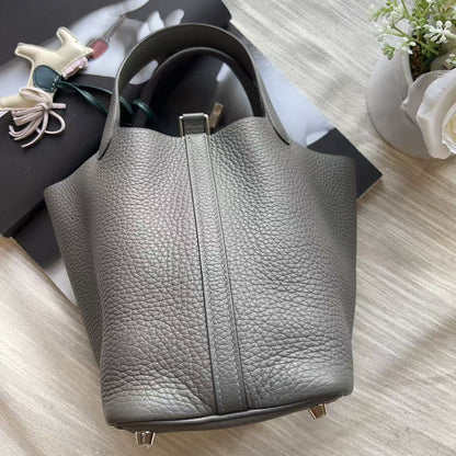 A gray pebbled leather handbag with double top handles, displayed on a light wood surface next to a black book and a small white flower pot.