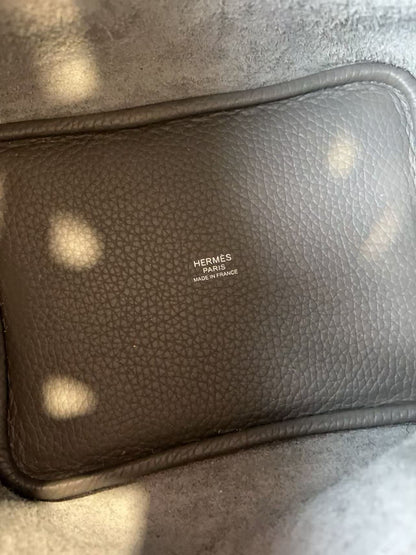 A close-up of a dark brown textured leather pouch with the HERMÈS PARIS MADE IN FRANCE logo embossed on the front, resting on a gray textured surface.