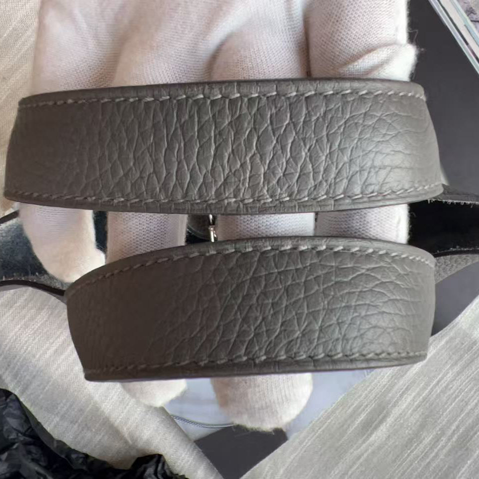 A close-up of a taupe brown textured leather belt being held by a white-gloved hand, showing the pebbled grain and stitching.