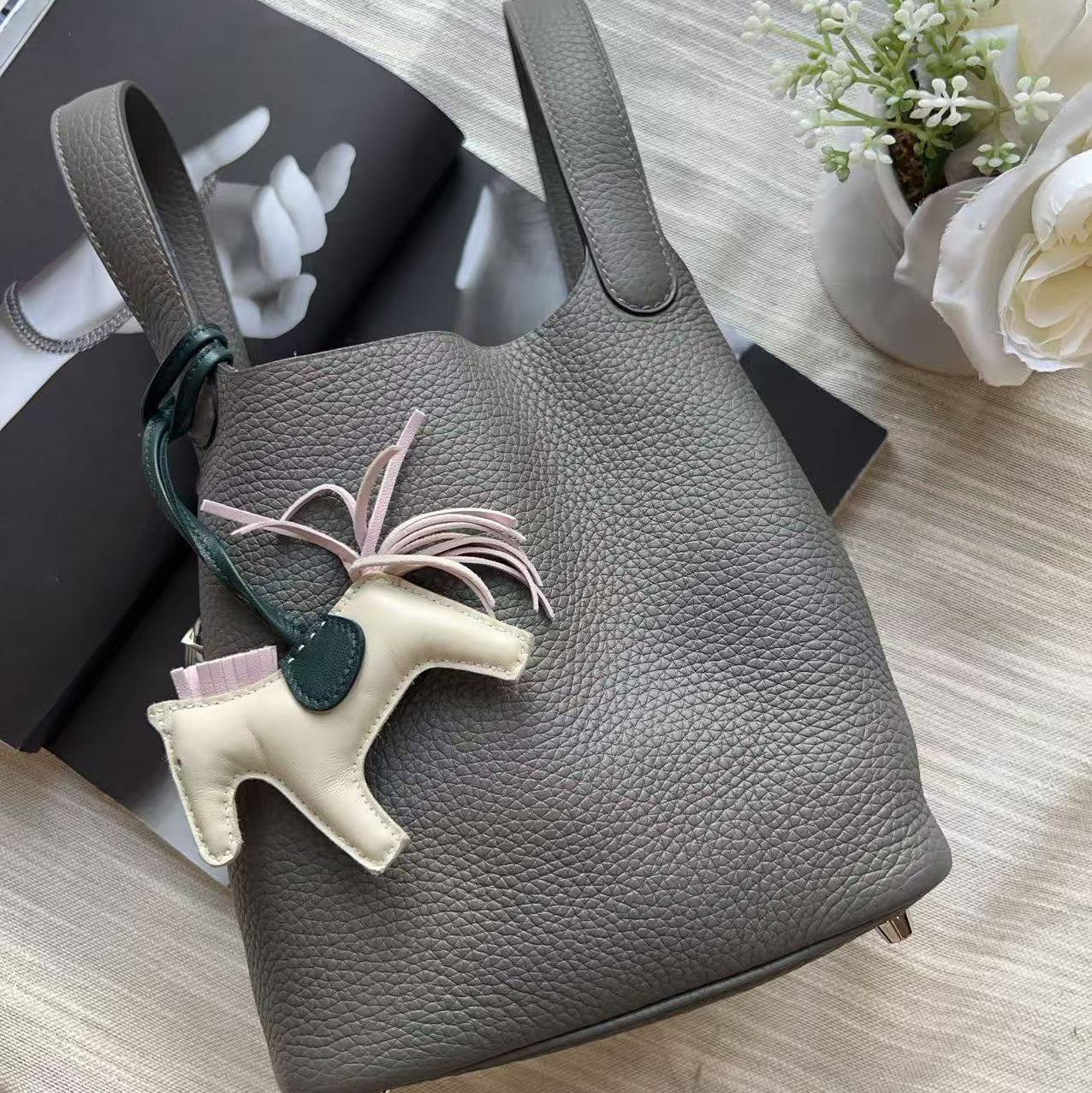 A gray pebbled leather bucket bag with a top handle, featuring a cream-colored horse charm with a dark green leather loop and a light pink ribbon, styled on a light beige fabric surface with books and white flowers.