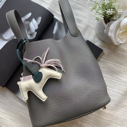 A gray pebbled leather bucket bag with a top handle, featuring a cream-colored horse charm with a dark green leather loop and a light pink ribbon, styled on a light beige fabric surface with books and white flowers.
