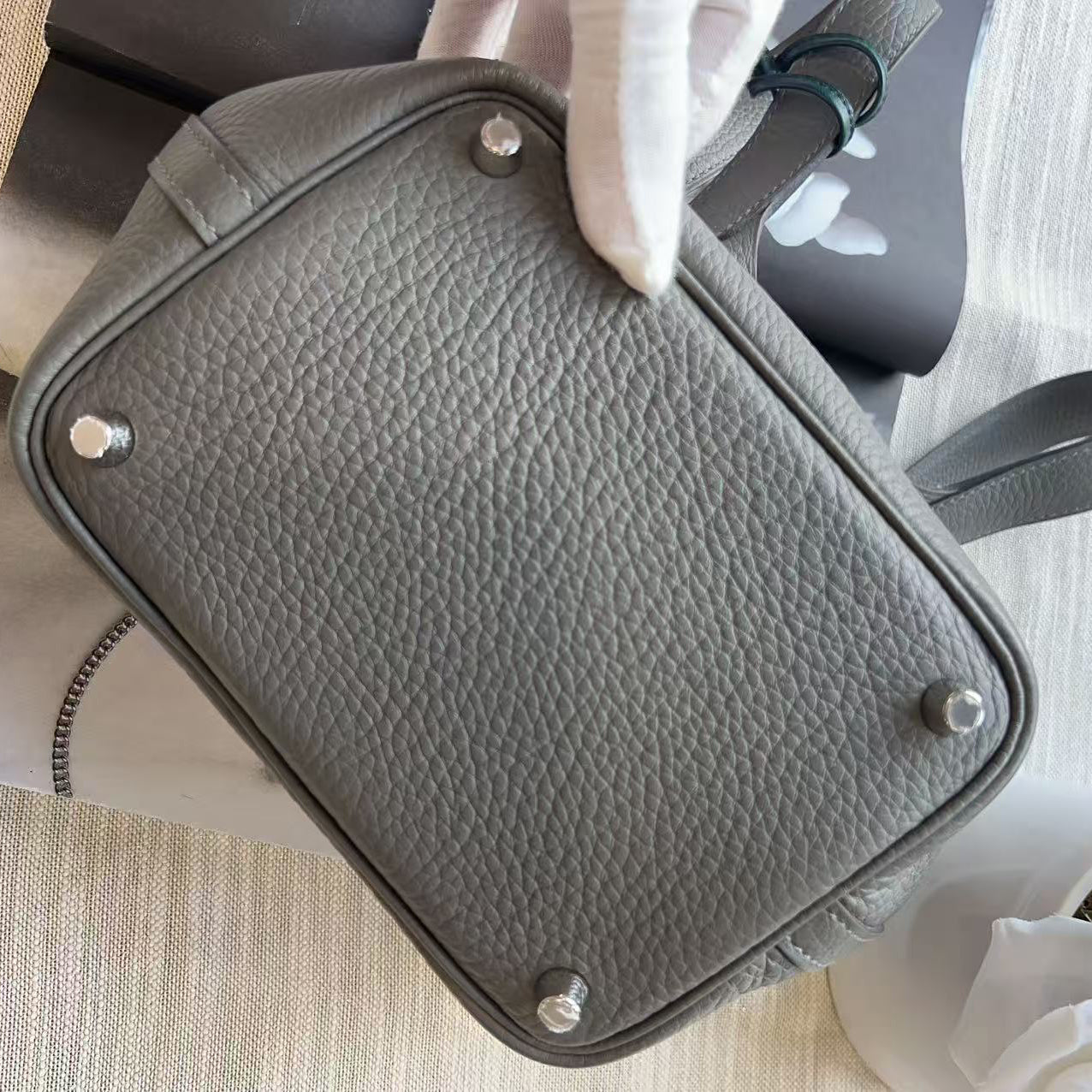 A gray pebbled leather handbag shown from the bottom, held by a hand wearing a white glove, with silver-tone metal studs on the base. The bag is photographed on a light beige textured surface with a small white flower in the corner.