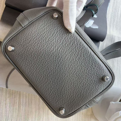A gray pebbled leather handbag shown from the bottom, held by a hand wearing a white glove, with silver-tone metal studs on the base. The bag is photographed on a light beige textured surface with a small white flower in the corner.