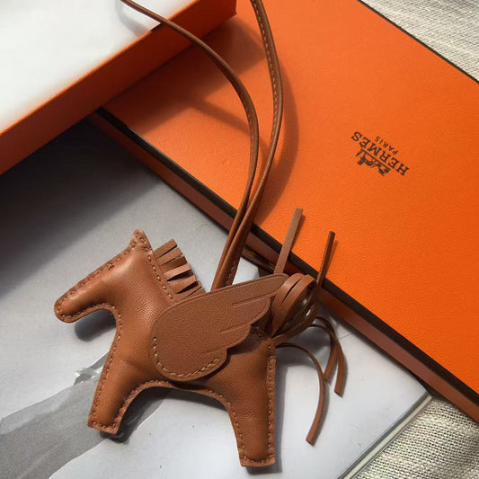 A brown leather winged horse charm with a matching strap, displayed next to an orange Hermès box on a gray surface.