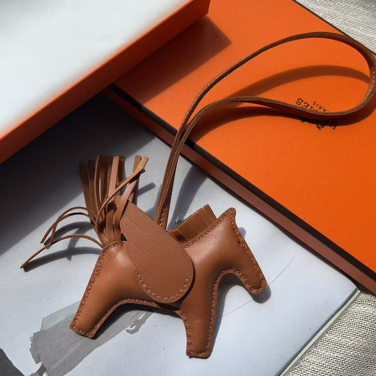 A tan leather horse-shaped keychain with a long strap, displayed on top of an orange box and a white card.