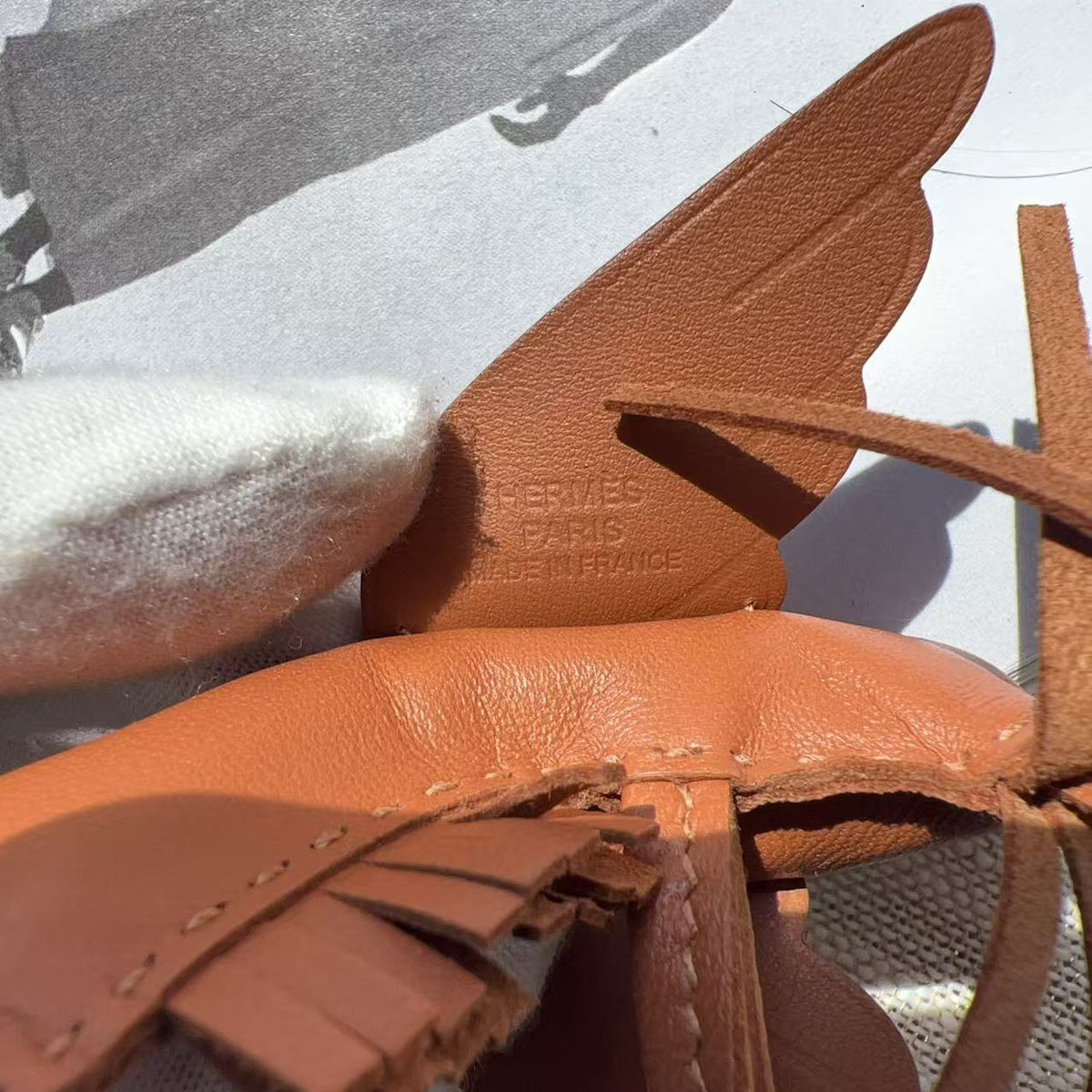 Close-up of a tan leather strap accessory with fringe and a wing-shaped leather tab, showing embossed Hermès Paris text on a light surface.
