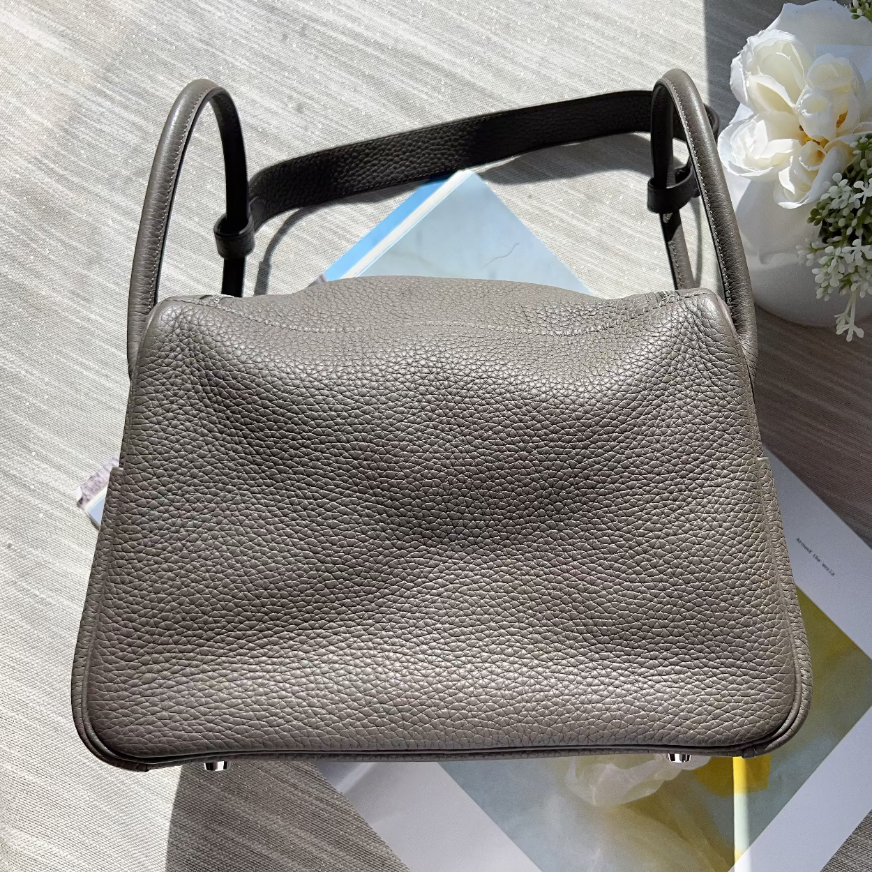 Small gray leather bag on a textured surface with flowers and a magazine in the background