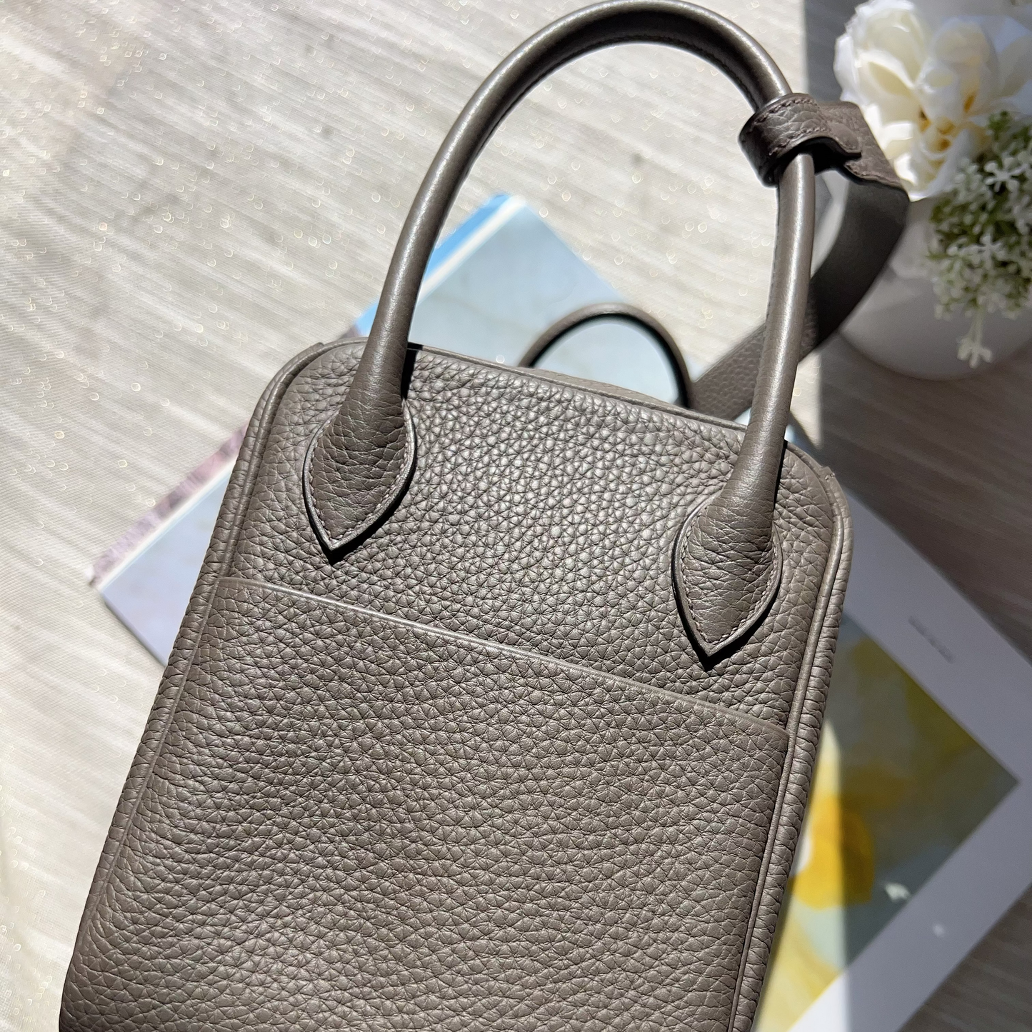 Gray leather handbag on a light wooden surface with a tablet and flowers.
