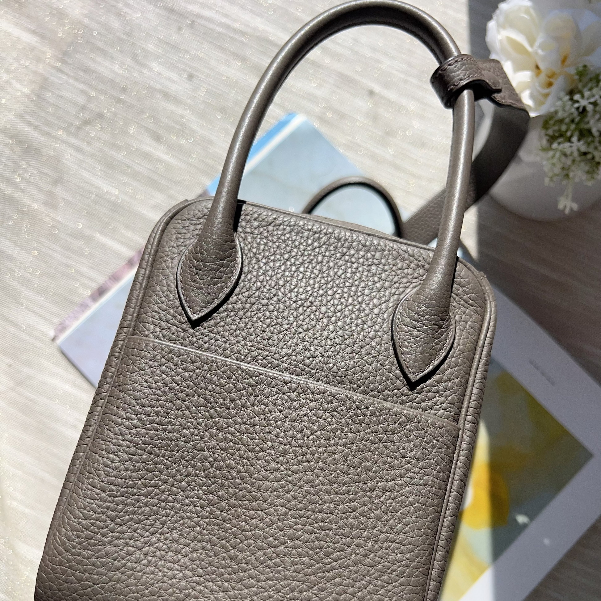 Gray leather handbag on a light wooden surface with a tablet and flowers.